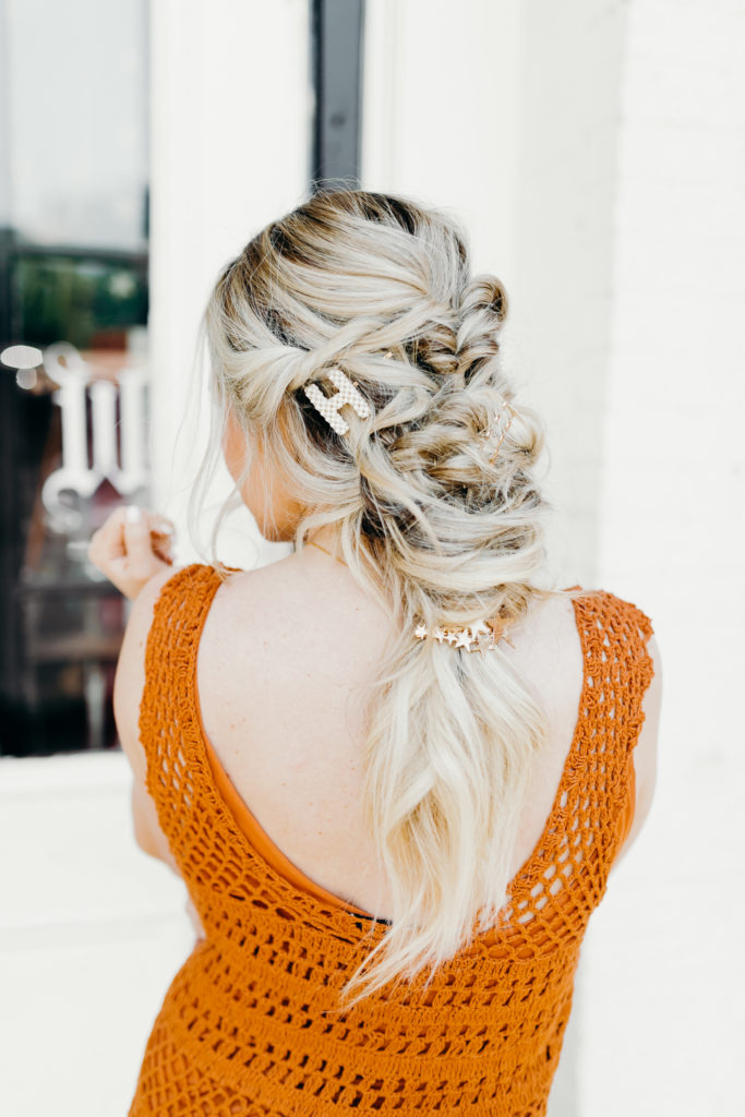 How to Wear Hair Clips by popular Nashville fashion blog, Nashville Wifestyles: image of a woman with a braided hairstyle and wearing a pearl letter H hair clip, gold bobby pins, and a star hair clip in her hair along with an orange crocheted maxi dress with fringe.